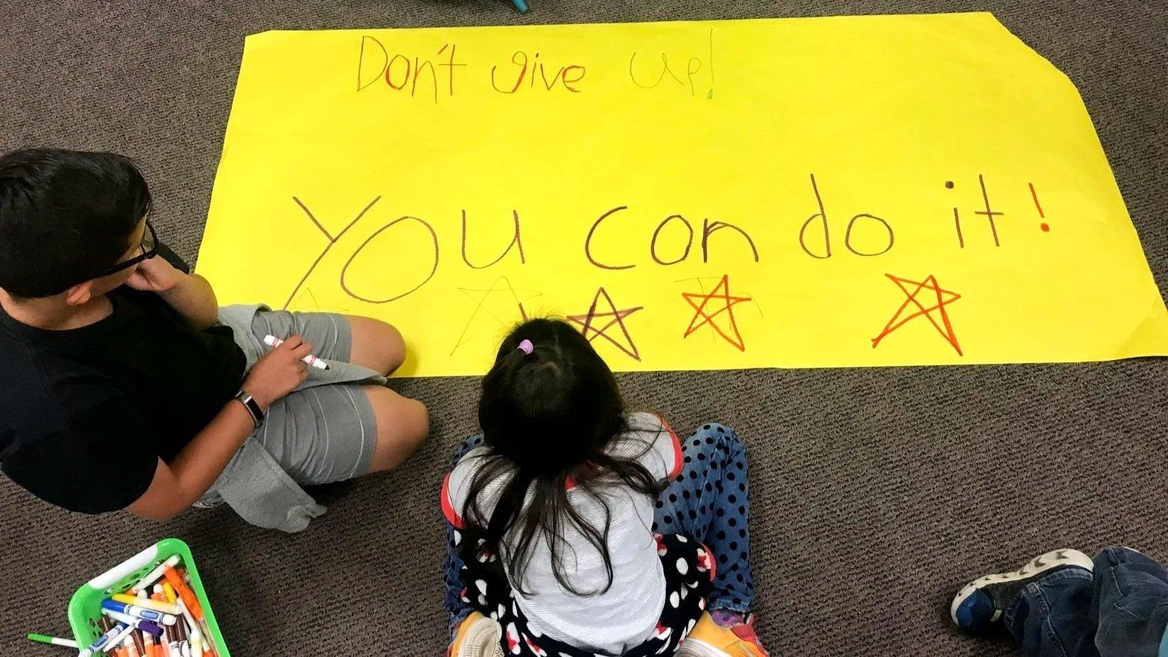 A large yellow poster on the floor with handwritten message 'Don't give up! YOU can do it!' in colorful marker, decorated with stars. Two children are sitting on the carpet, one girl with long dark hair and a white shirt with red and black dots, and a boy with glasses and a black shirt. A basket of markers is nearby.