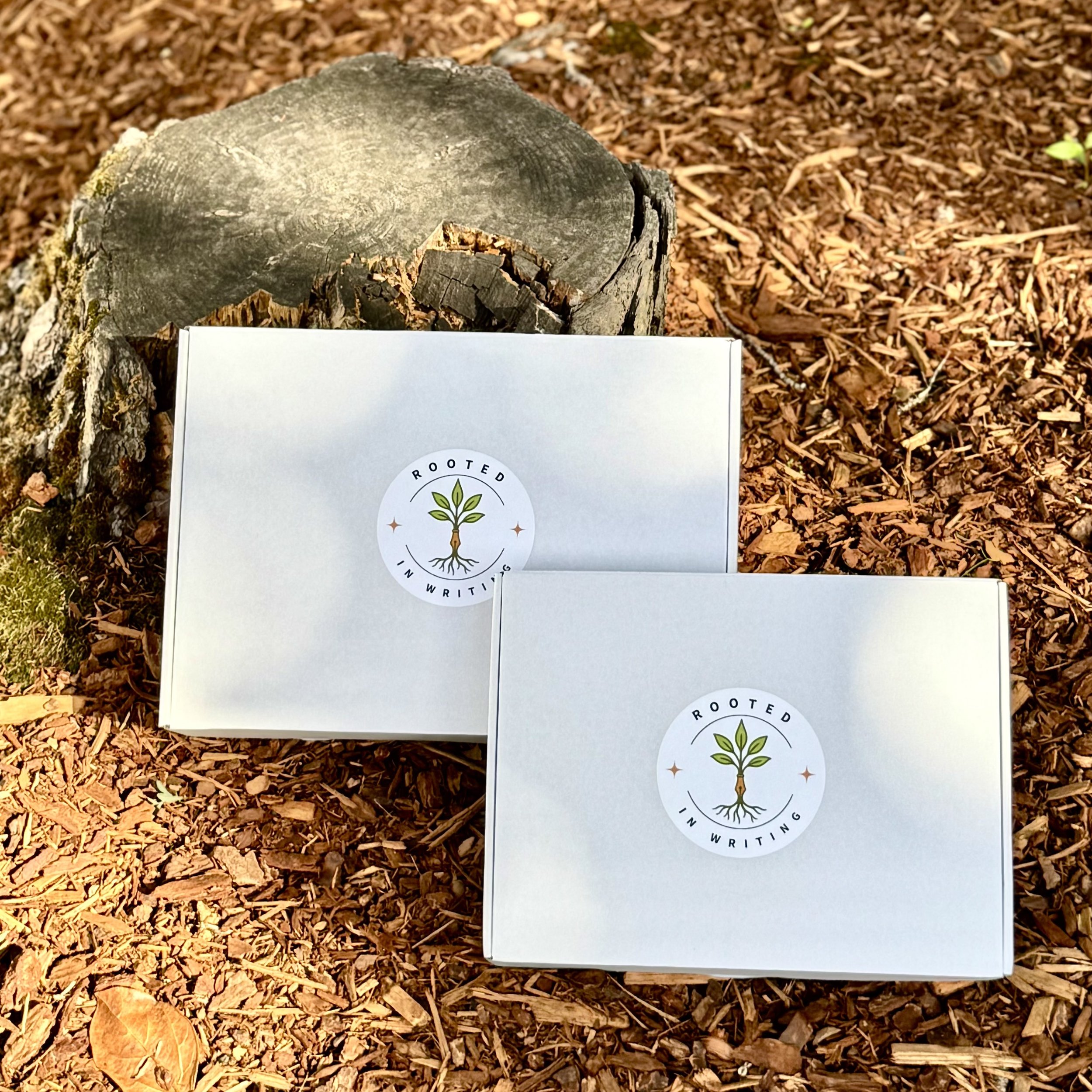 Two white boxes with the logo 'Rooted In Writing' featuring a tree with roots and leaves, placed outdoors on brown bark mulch next to a tree stump.